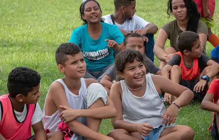 Children sitting in a field (grass), smiling as part of One Social Investment's strategies, achieving social investment through strategic philanthropy for transformative initiatives.