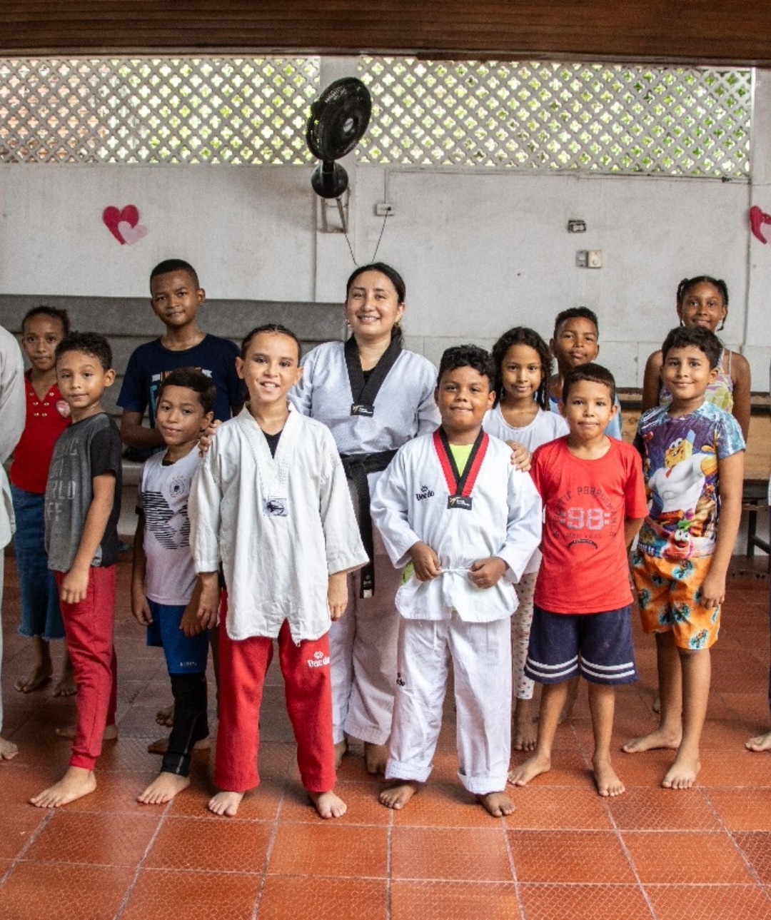 Grupo de jóvenes, en su mayoría niños, parados en fila. En el centro del grupo, una joven viste un uniforme blanco de karate, al igual que algunos de los otros niños. Todos están sonriendo y mirando a la cámara, transmitiendo una sensación de alegría y unión. El fondo es una pared blanca simple, lo que permite que los sujetos destaquen. La composición de la imagen es excelente, con colores vibrantes, y captura de manera conmovedora la alegría de la juventud y la camaradería del grupo. Esta imagen refleja el impacto positivo de los proyectos de One Inversión Social, que promueven la integración y el desarrollo de habilidades a través de actividades enriquecedoras.
