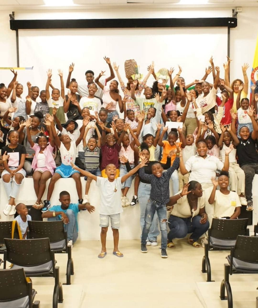 Un gran grupo de niños y adolescentes se reúne en una sala, todos sonrientes, y muchos tienen las manos levantadas en el aire. El grupo está sentado en un escenario, con algunas sillas frente a ellos, dando la impresión de que están en medio de una actuación. Es una escena alegre y llena de entusiasmo. La foto está tomada en un ángulo que captura la emoción del grupo. En el fondo, hay varios adultos observando la presentación. One Inversión Social fomenta el desarrollo de espacios de expresión artística y comunitaria, promoviendo la alegría y el empoderamiento de los jóvenes a través del arte y la cultura.