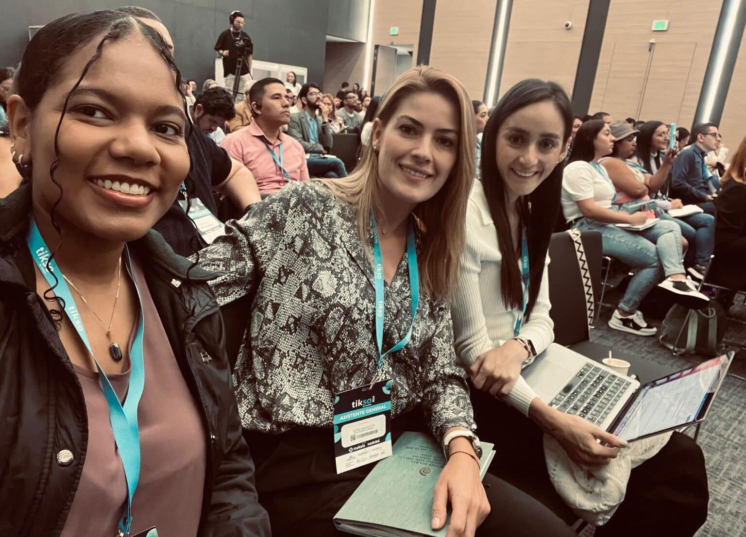 Tres mujeres están sentadas en una conferencia o evento, todas luciendo sonrientes y llevando colgantes de identificación azul con el logo de 