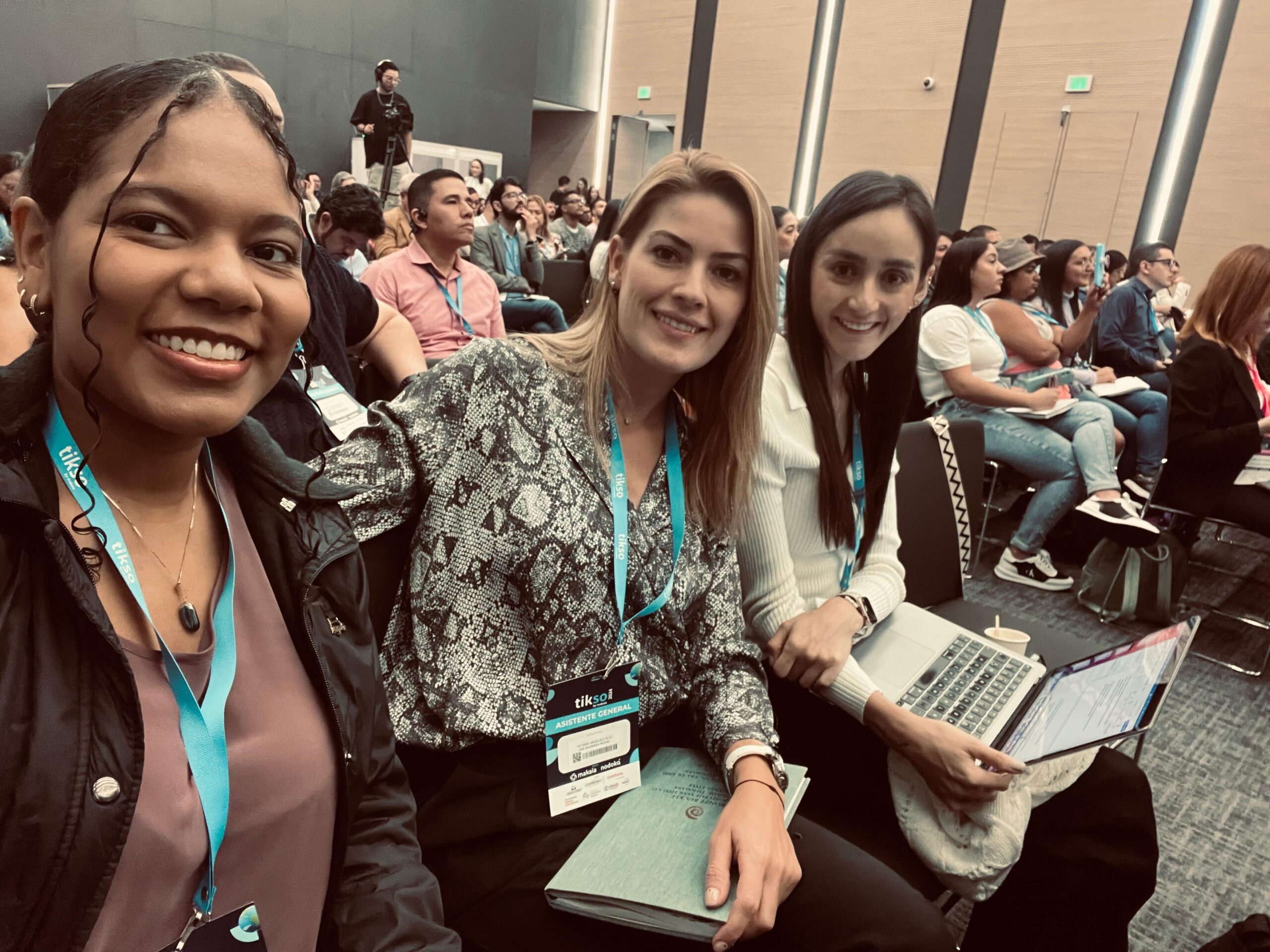 Three women are sitting at a conference or event, all smiling and wearing blue identification badges with the 